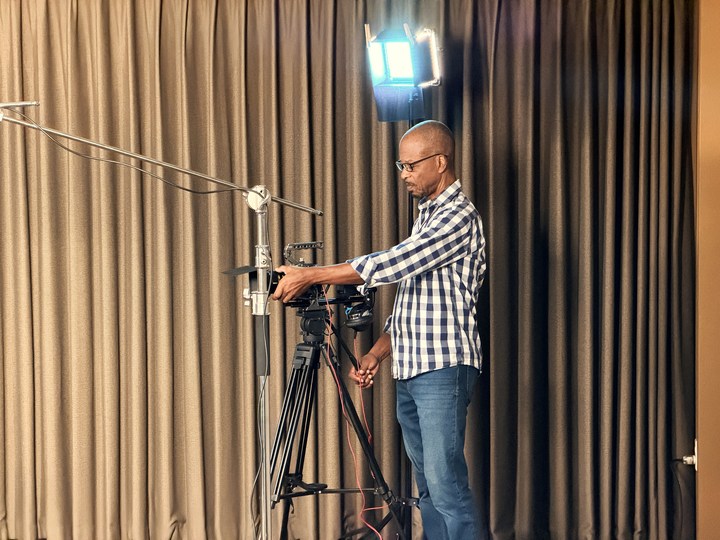 A participant adjusting a tripod-mounted camera beside the rehearsal backdrop