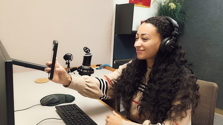 A person wearing headphones and holding a phone beside the Podcast Room A microphone